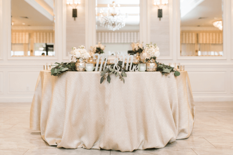 An elegantly decorated sweetheart table with a white tablecloth is set up in a luxurious venue. The table features floral arrangements, greenery, and a "Mr & Mrs" sign. In the background, there are mirrors, chandeliers, and wall sconces adding to the sophisticated ambiance.