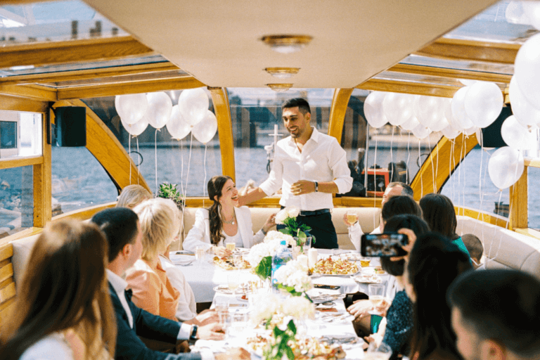 A group of people are seated around a decorated table on a boat. The table has food, flowers, and drinks. A man in a white shirt is standing, speaking while raising a glass, with balloons in the background. The setting is bright and festive with a seaside view.