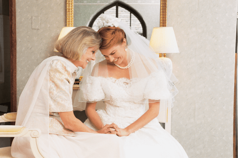 A bride and an older woman, possibly her mother or grandmother, share a tender moment. Both are seated and leaning their heads together with their eyes closed, smiling. The bride wears a white dress, veil, and pearls, while the older woman wears a light-coloured dress.