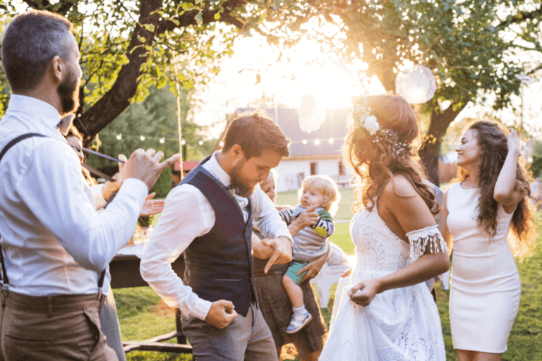 A joyful outdoor wedding celebration with guests dancing and clapping. The bride, in a white dress and floral headpiece, dances with a young child in her arms. The sun sets in the background, casting a warm glow over the festive scene.
