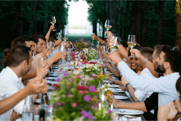 A large group of people seated at a long, outdoor dining table adorned with colorful flowers and tableware, raising their glasses in a celebratory toast. The setting is surrounded by trees, creating a festive and enchanting atmosphere.
