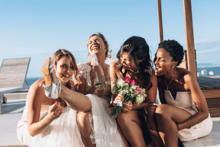 A joyful bride and her three bridesmaids sit laughing and smiling on a wooden deck by the ocean. The bride holds a drink and one bridesmaid playfully lifts her leg, while another holds a bouquet of flowers. They are all dressed in light-coloured gowns.