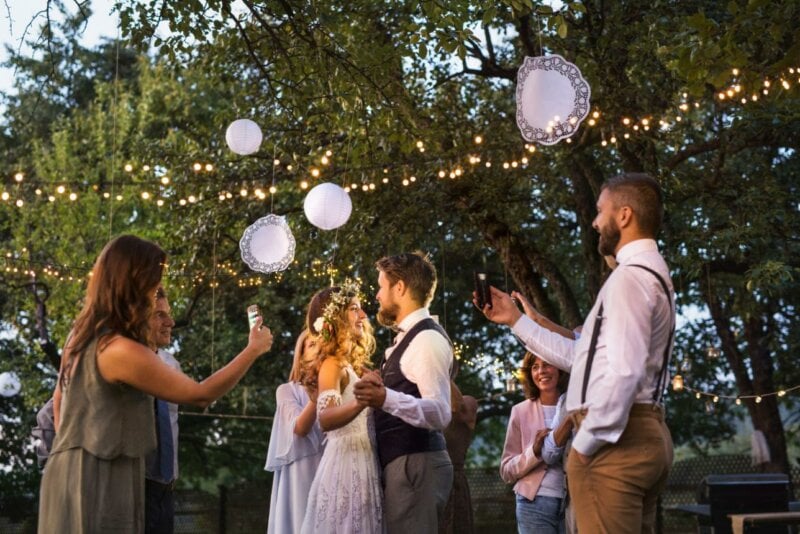 Couple de mariés sous un arbre décoré de guirlandes lumineuses et invités