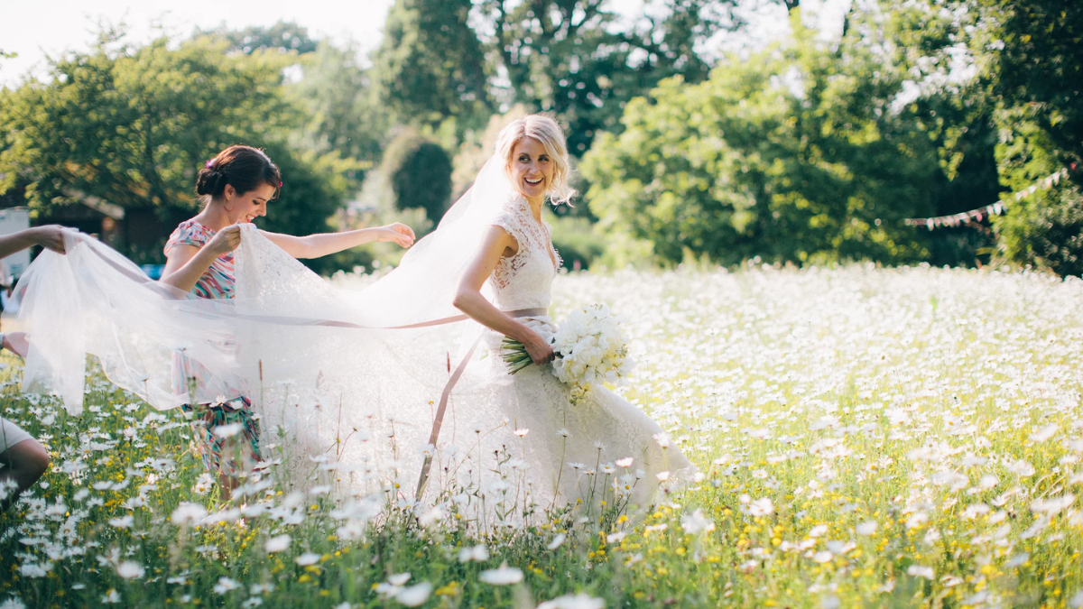 Smiling bride in a field with bridesmaids holding their dress