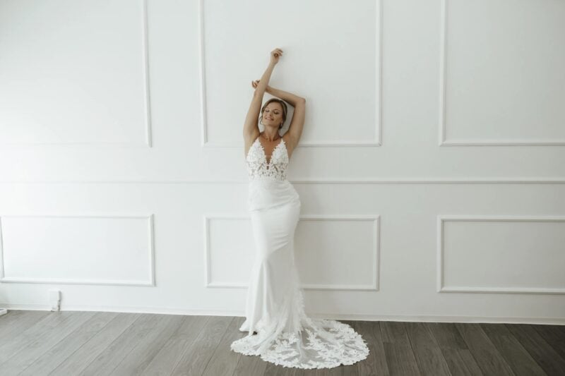 Braut in modernem, figurbetontem Brautkleid mit Spitzenoberteil vor weißer Wand
