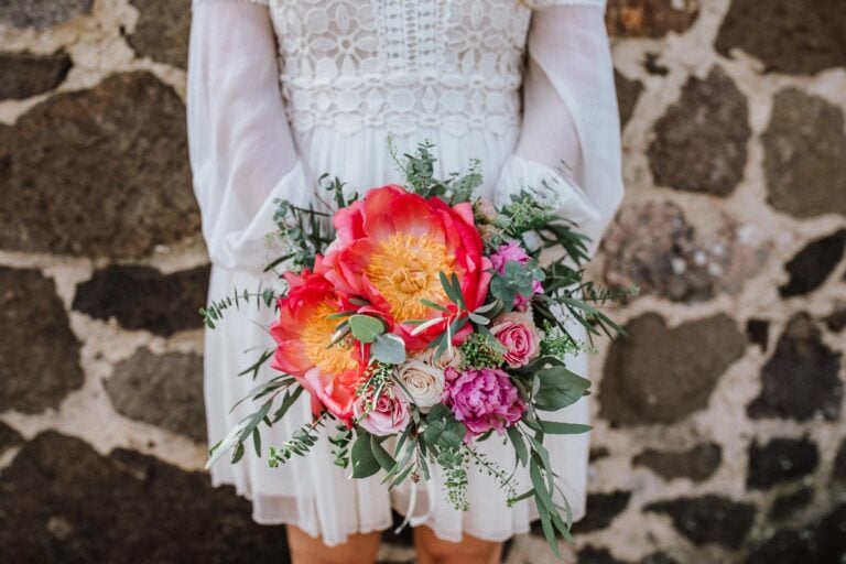 Braut hält farbenfrohen Brautstrauß mit Pfingstrosen, Rosen und Eukalyptus vor rustikaler Steinwand