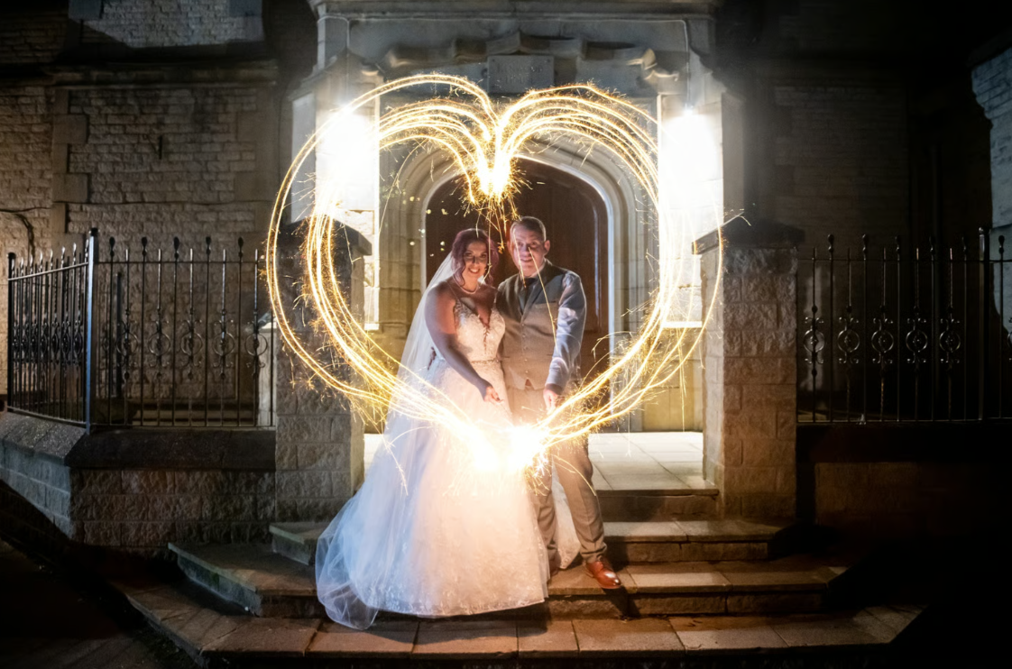 Newly wedded couple outside on wedding venue steps using sparklers to make a heart shape