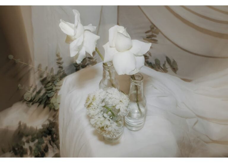 Minimalistische Hochzeitsdeko mit weißen Rosen und zarten Blüten in Glasvasen auf hellem Stoff arrangiert