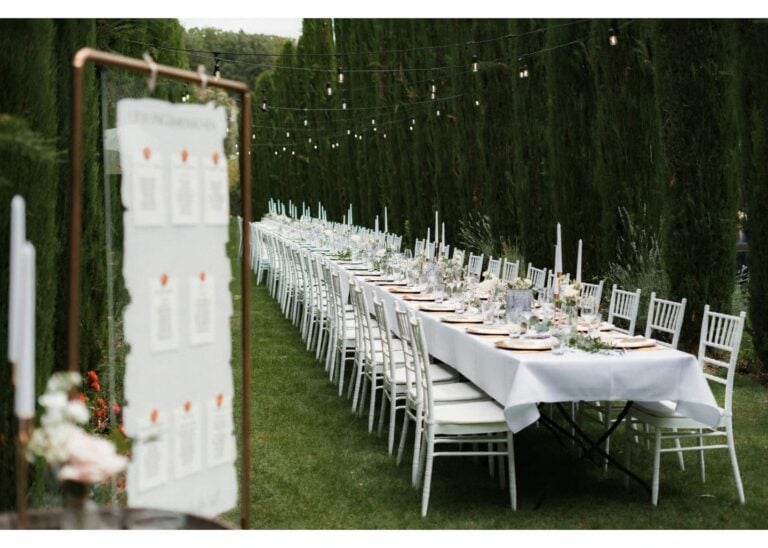 Lange Hochzeitstafel im Freien mit weißen Stühlen und Lichterketten zwischen hohen Hecken