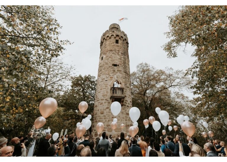 Hochzeitsgesellschaft mit Ballons vor einem historischen Turm, während das Brautpaar vom Balkon schaut