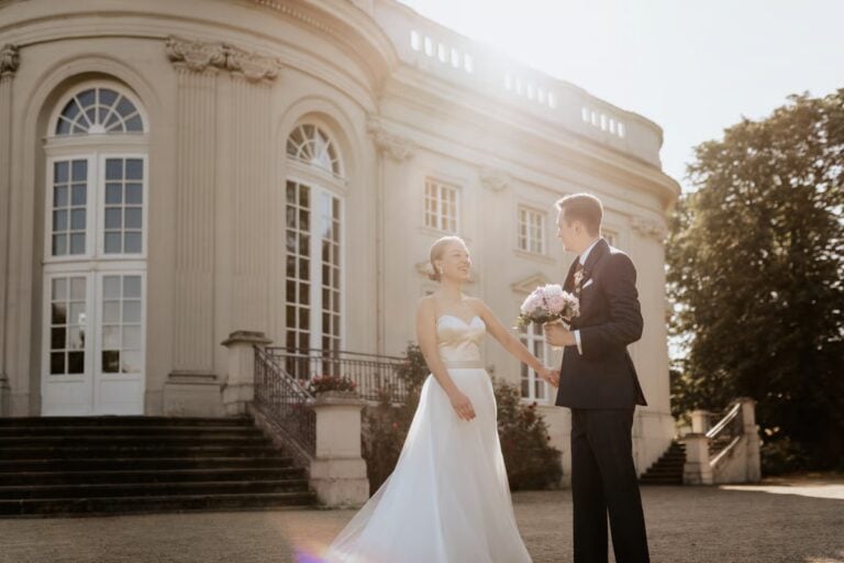 Braut und Bräutigam stehen vor einer eleganten Hochzeitslocation und halten sich lächelnd an den Händen