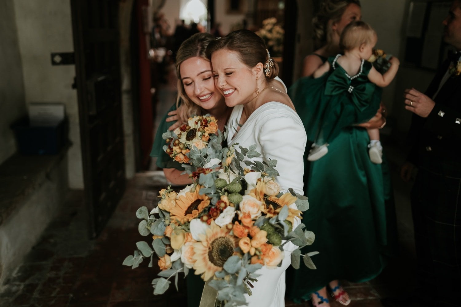 a bride hugging her bridesmaid as she holds an oversized bouquet of peach and white flowers with greenery.