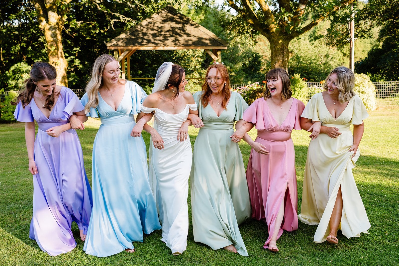 a bride arm in arm with her bridesmaids who are all wearing different pastel-coloured dresses