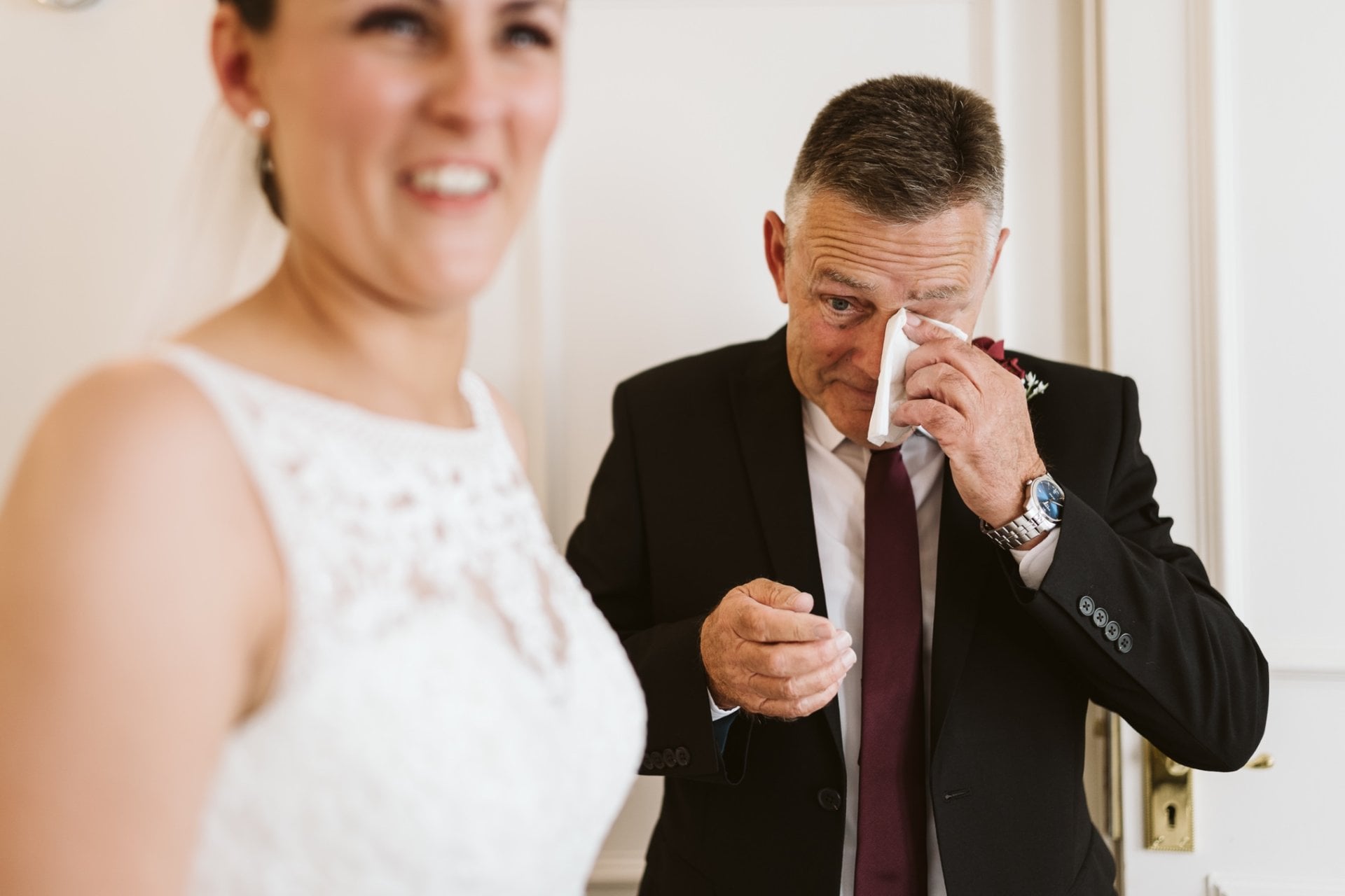 a father of the bride wipes a tear from his eye as his daughter, the bride, smiles in the foreground
