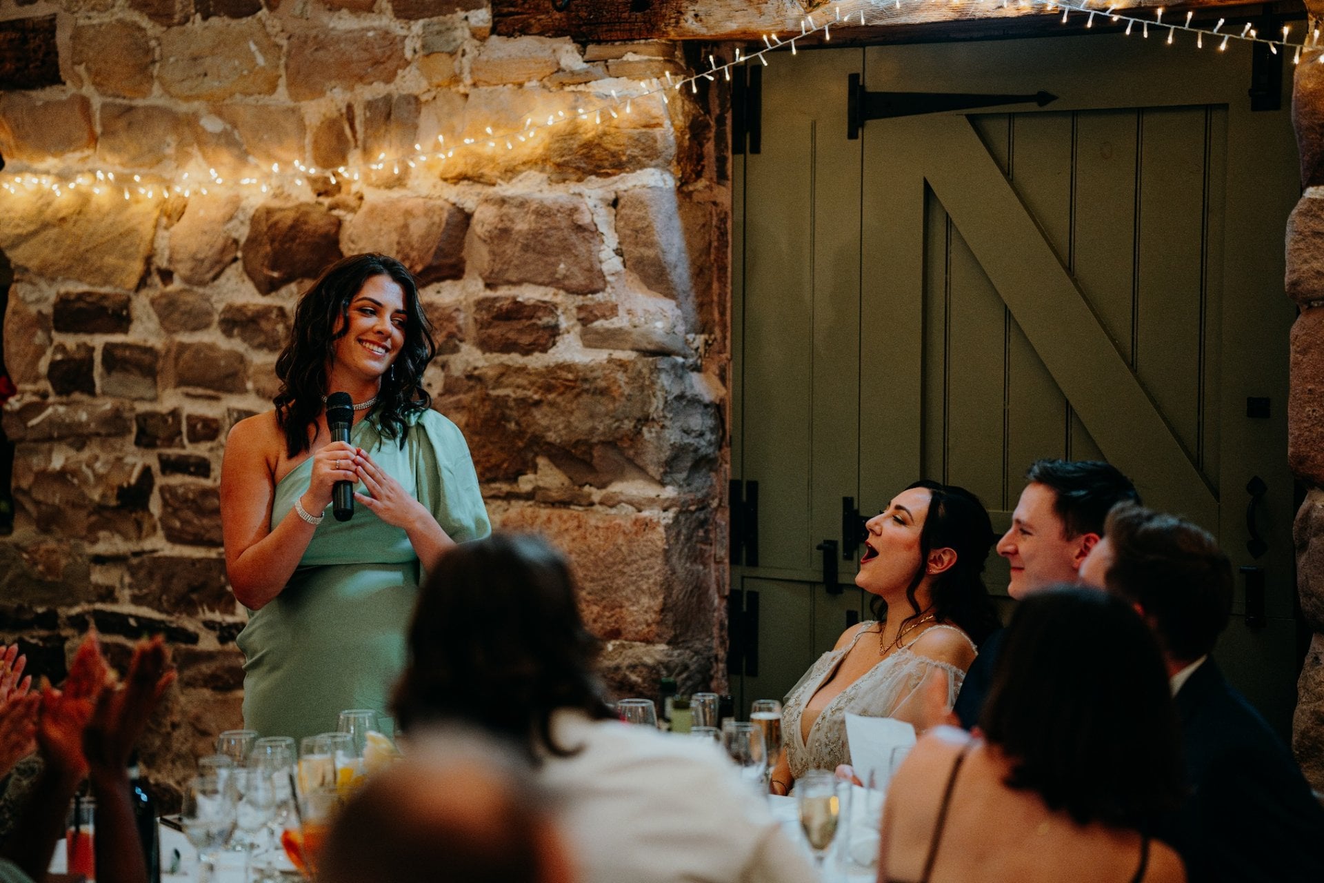 a maid of honour in a green dress giving a wedding speech as the bride laughs