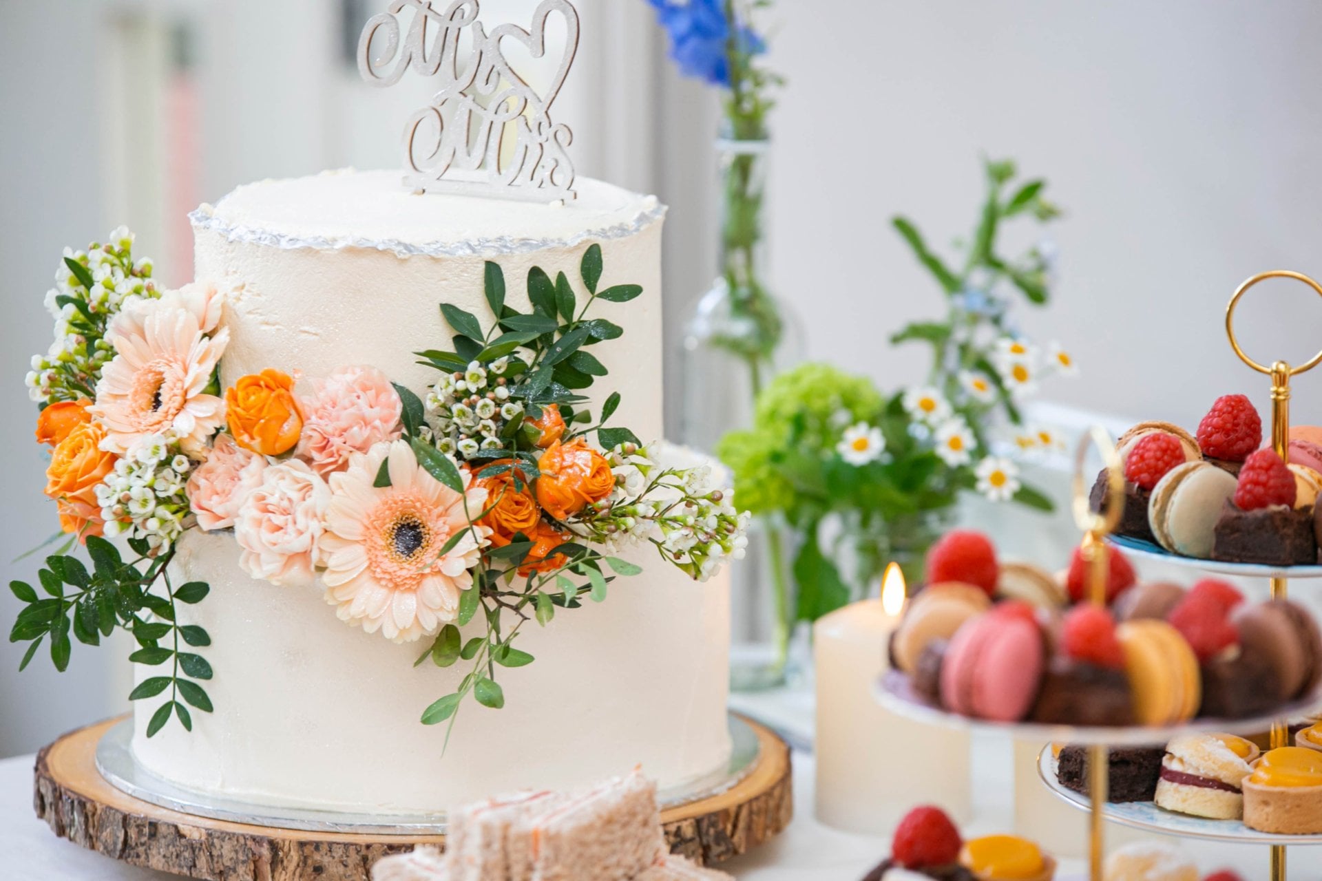 a white two-tier wedding cake decorated with fresh flowers on a table with a cake stand filled with colourful macarons