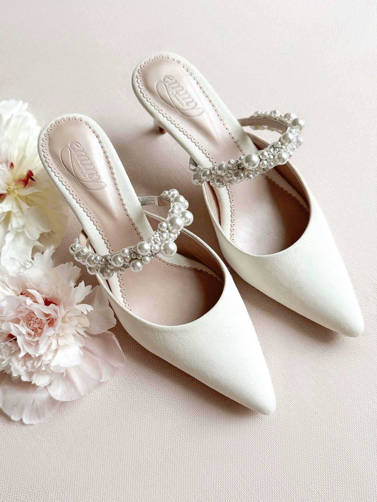 a pair of pointy ivory mules with crystal strap along the top, they have been placed next to a bouquet