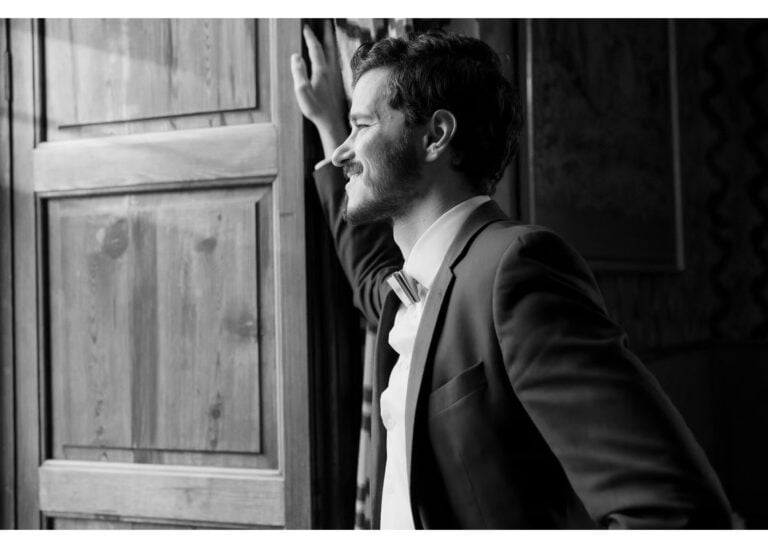 Bräutigam lehnt lächelnd am Fenster und blickt nach draußen im Hochzeitsanzug