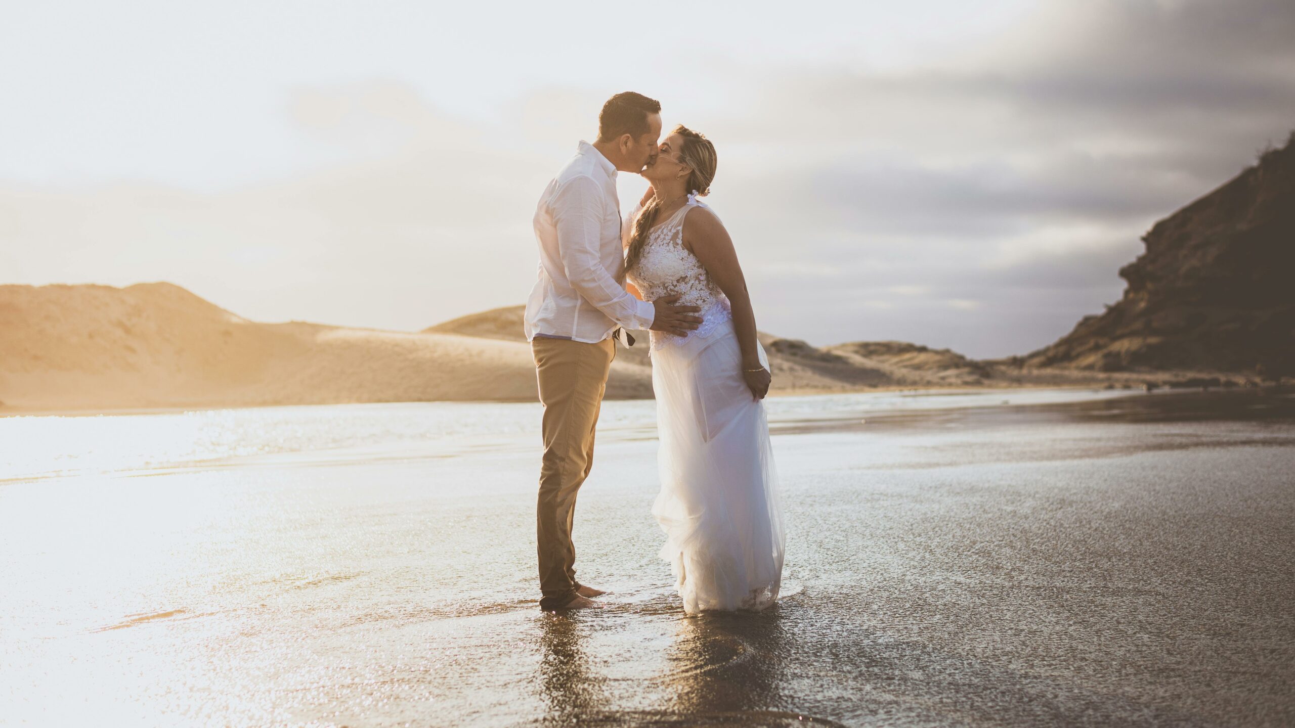 Newlyweds in wedding attire kissing on the shore in the waves
