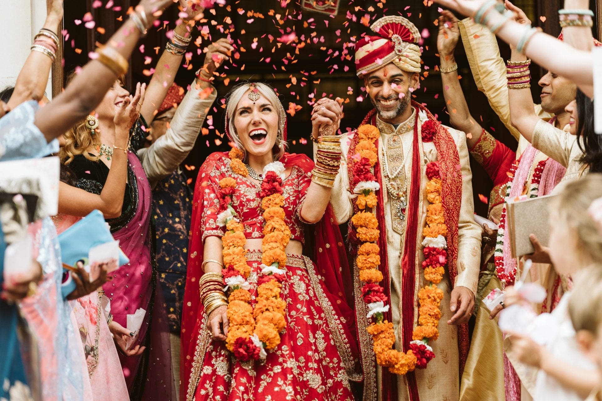 a bride and Indian groom leave their wedding ceremony in traditional Hindi dress and floral garlands as guests throw petal confetti