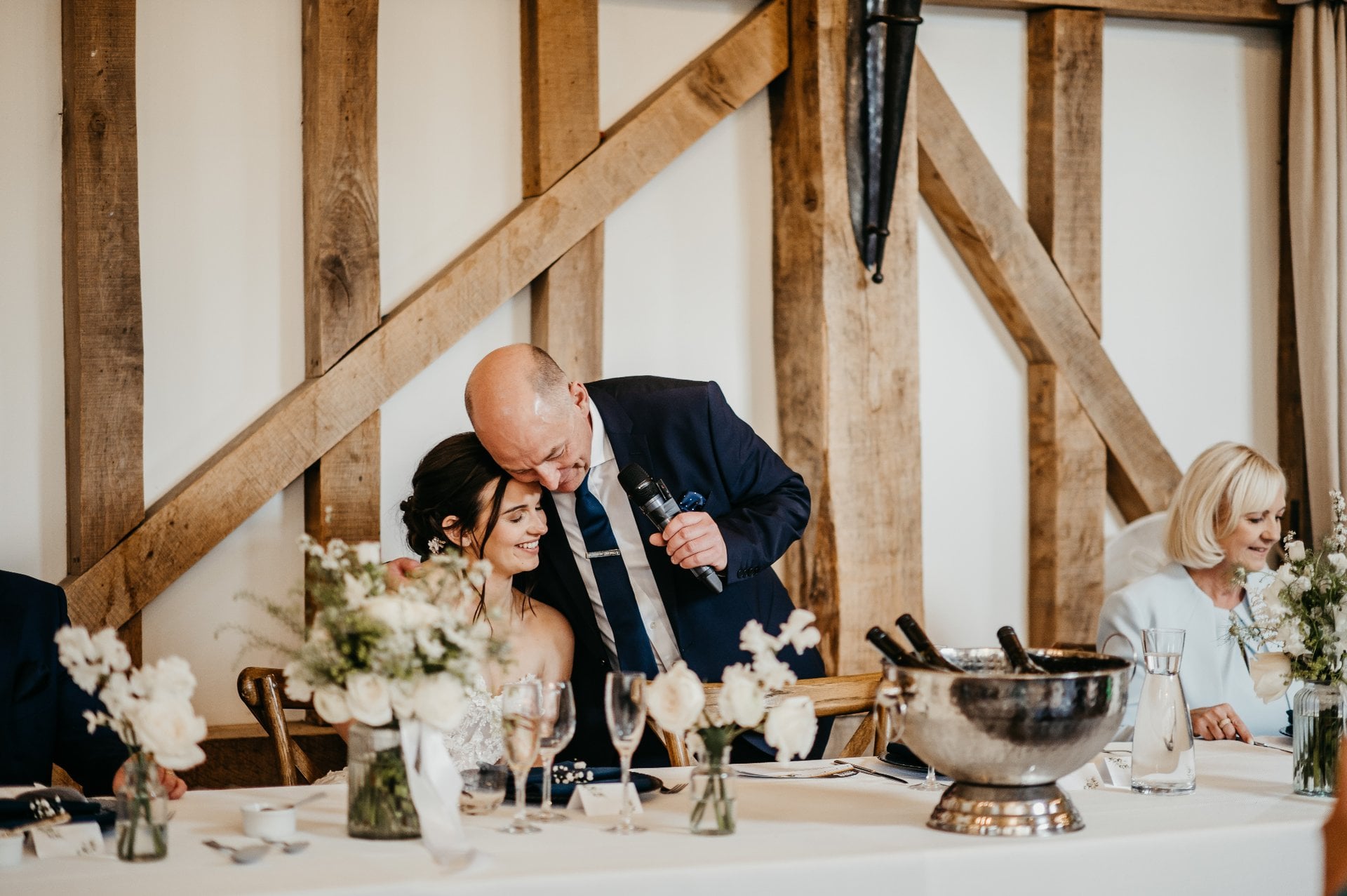 a father of the bride holds a microphone and hugs his daughter at the top table