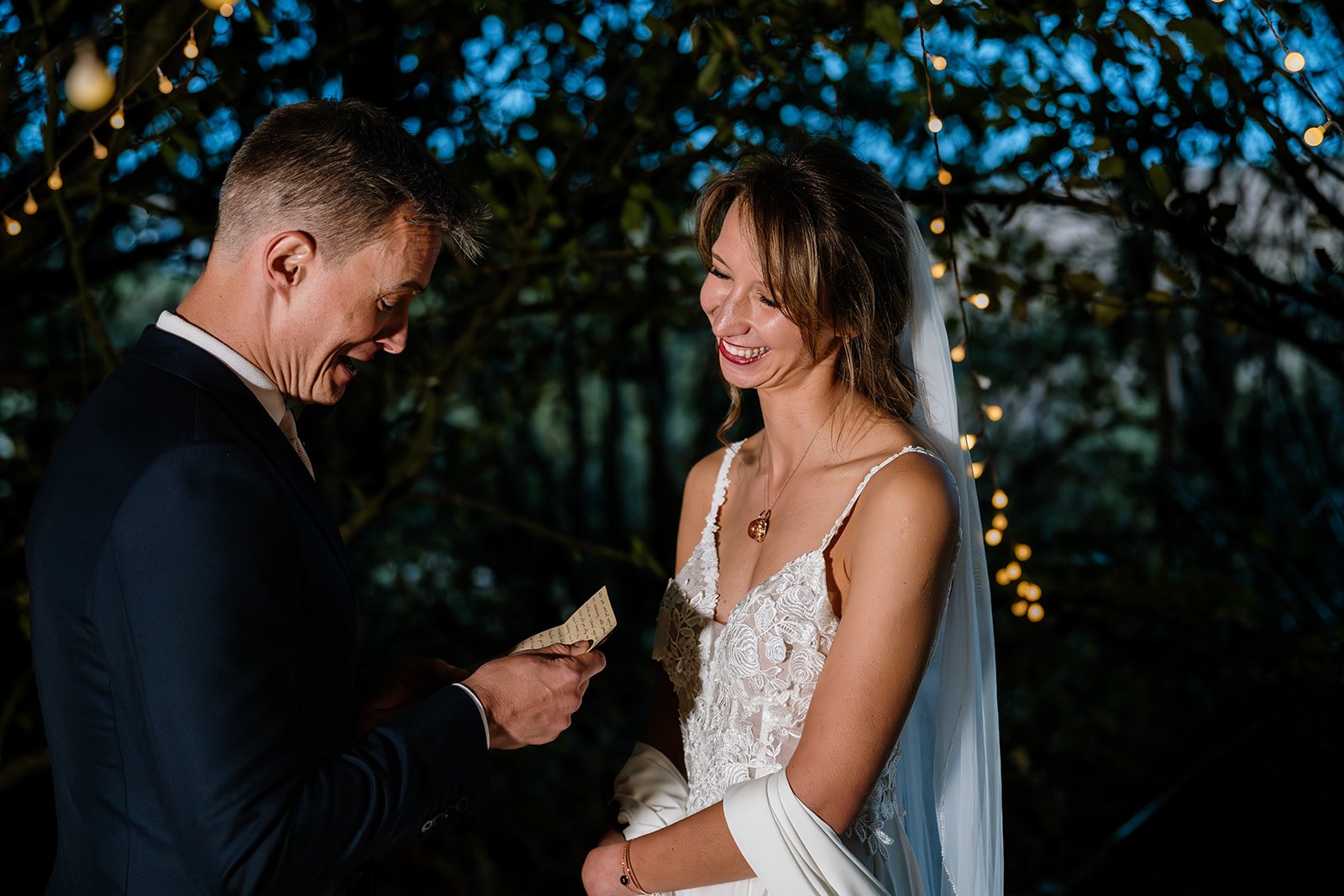 a groom reading from a sheet of paper to his bride as she smiles and holds his hand