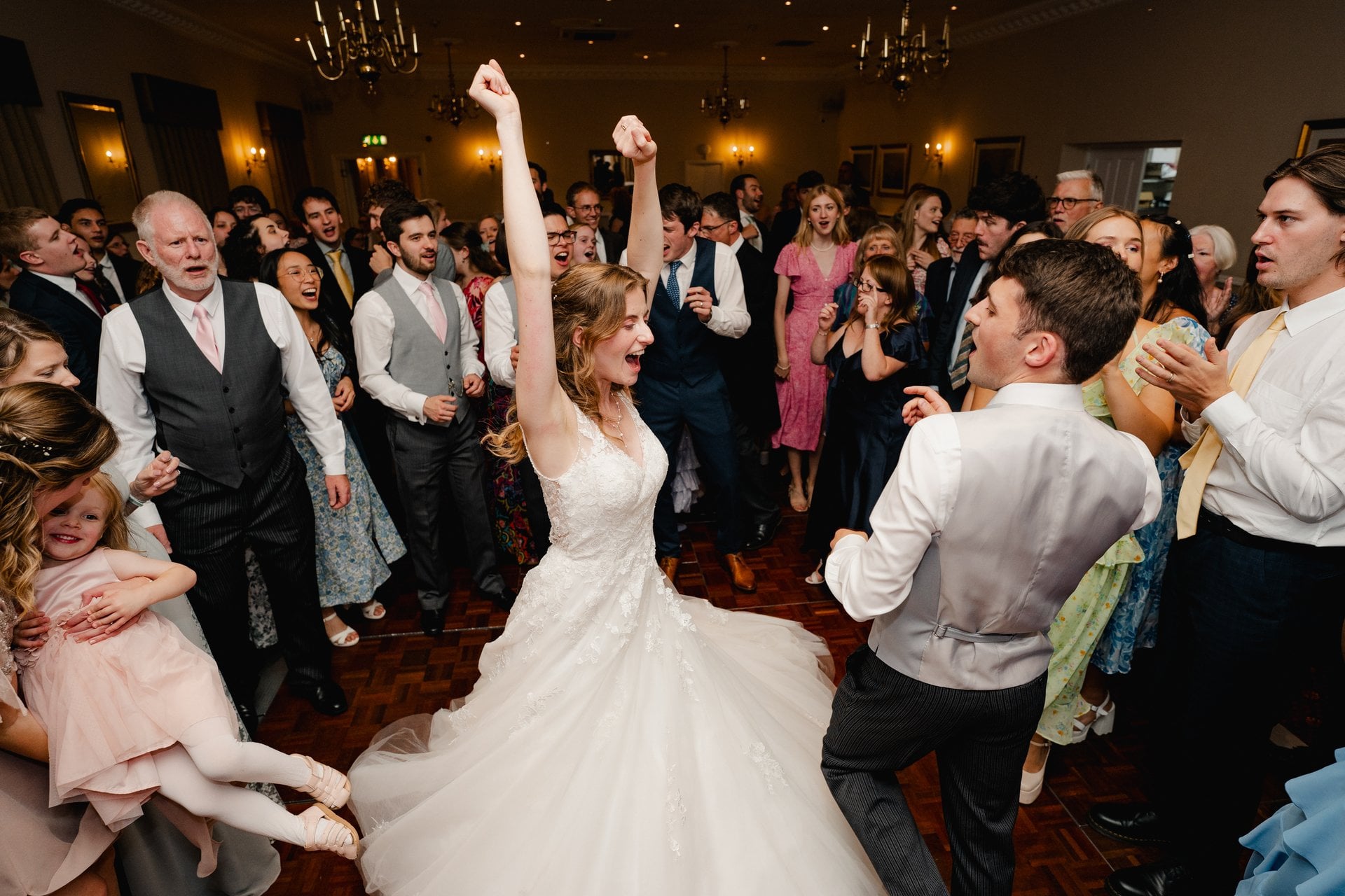 a bride and groom dancing as their guests surround them, the bride has her arms in the air and is singing along