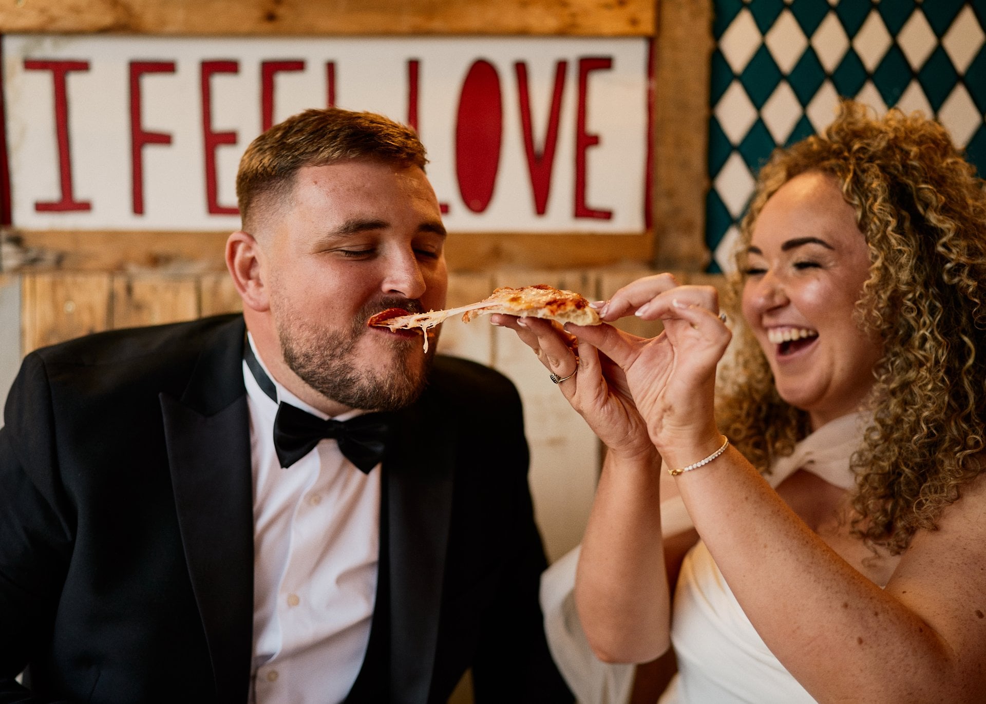 a bride with curly hair feeding her groom pizza and laughing. in the background is a sign that reads, 'i feel love'