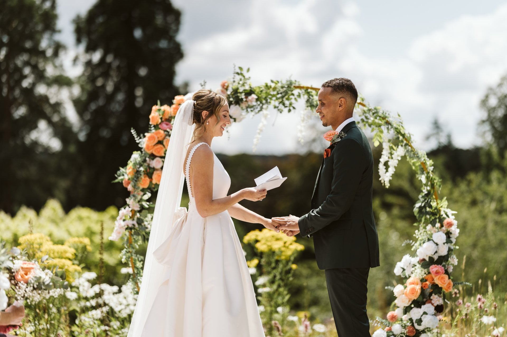 a bride standing in front of her groom at their outdoor wedding ceremony reading her vows from a piece of paper and holding his hand.