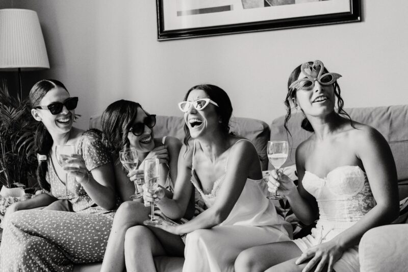 Braut und Freundinnen lachen gemeinsam mit Sonnenbrillen und Sekt auf dem Sofa